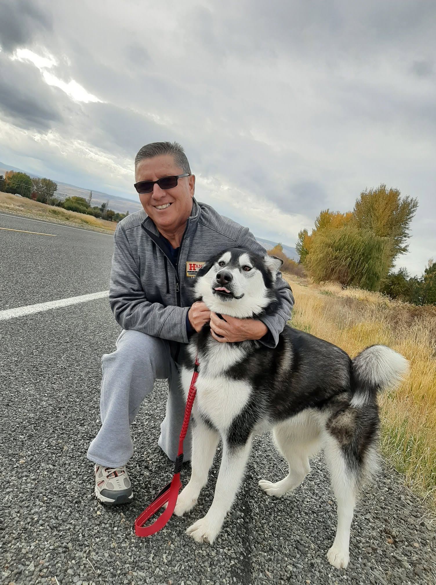 Man in sunglasses and gray jacket posing with black and white husky dog on road