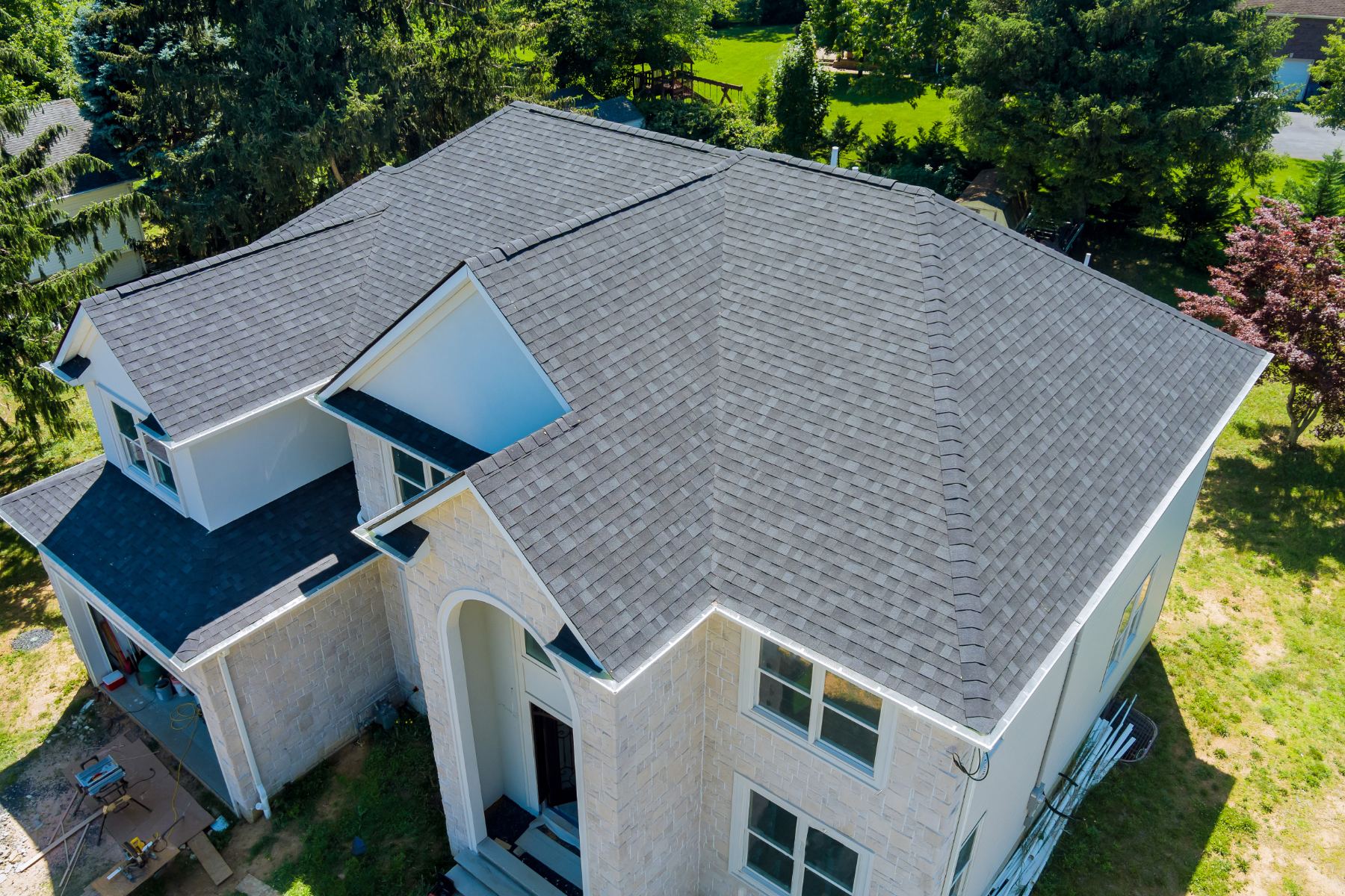 Aerial view of residential house with gray asphalt shingle roof and white brick exterior surrounded by green lawn