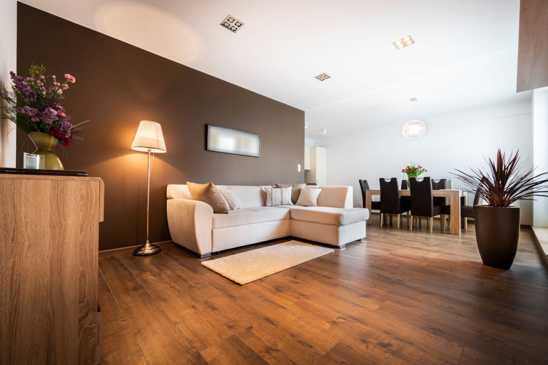 Modern open-plan living room with beige sectional sofa, brown accent wall, wooden furniture and dining area